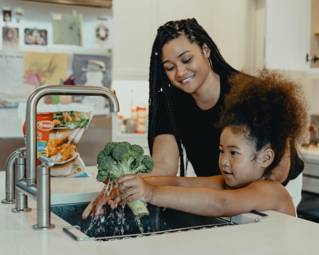 Woman and young daughter washing broccoli in sink.